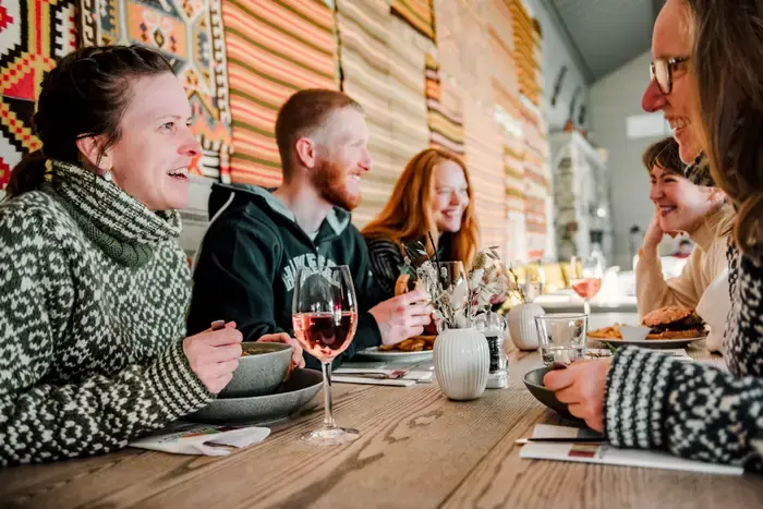 Venner et lunsj ved eit langbord framfor fargerike, vovne åklede